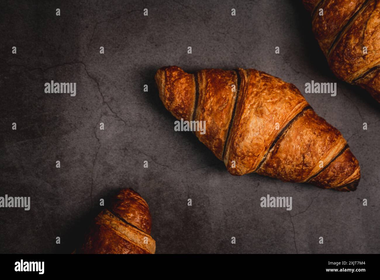 Drei frisch gebackene Croissants sitzen auf einer dunkelgrauen Arbeitsplatte, bereit zum Verzehr. Ihre goldbraune Kruste glitzert unter dem weichen Licht und lädt ein Stockfoto