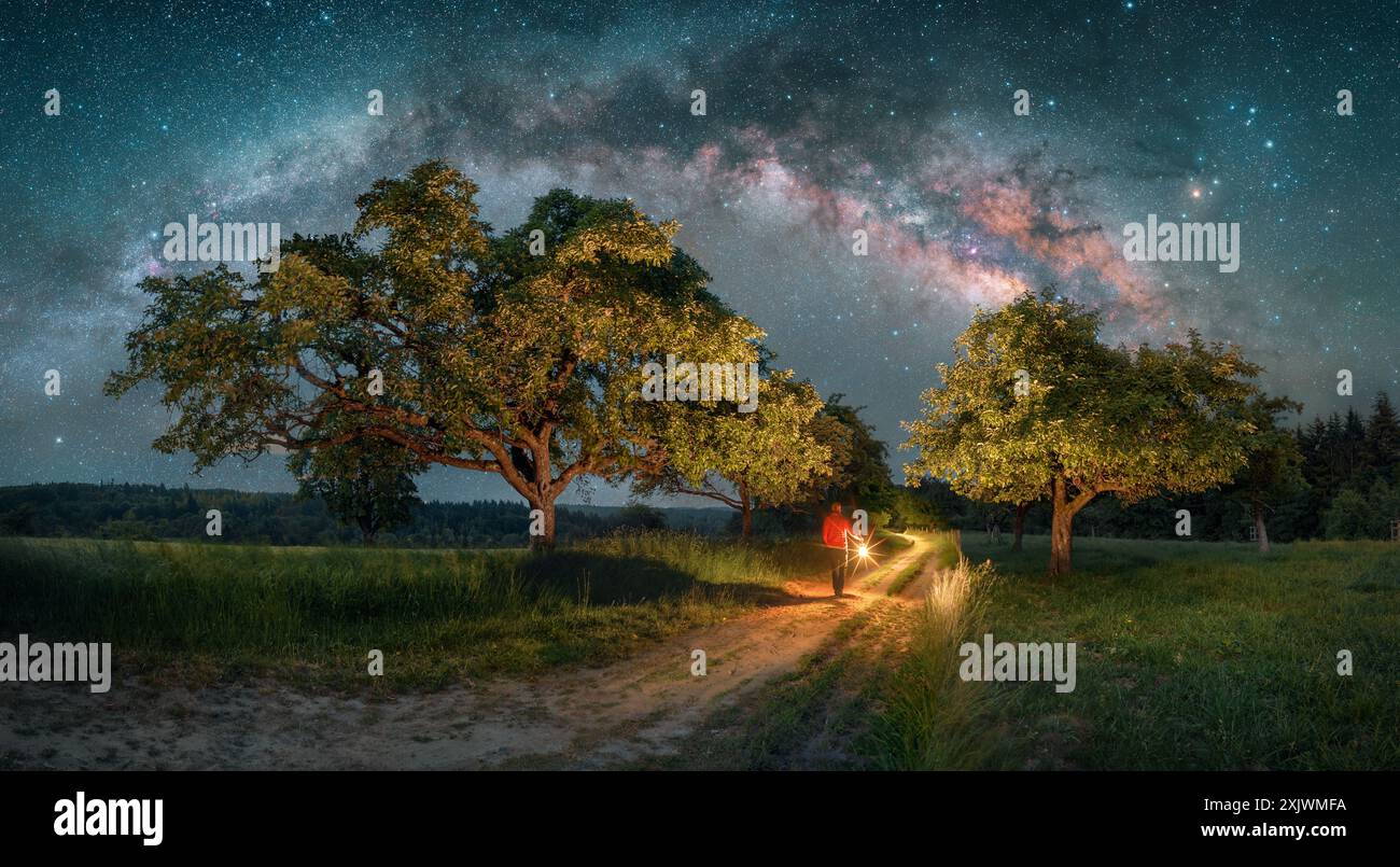 Mann mit Laterne, der unter dem Himmelsbogen der Milchstraße läuft. Eine ländliche Landschaft mit Bäumen auf einer Wiese, die einen Feldweg umgibt Stockfoto