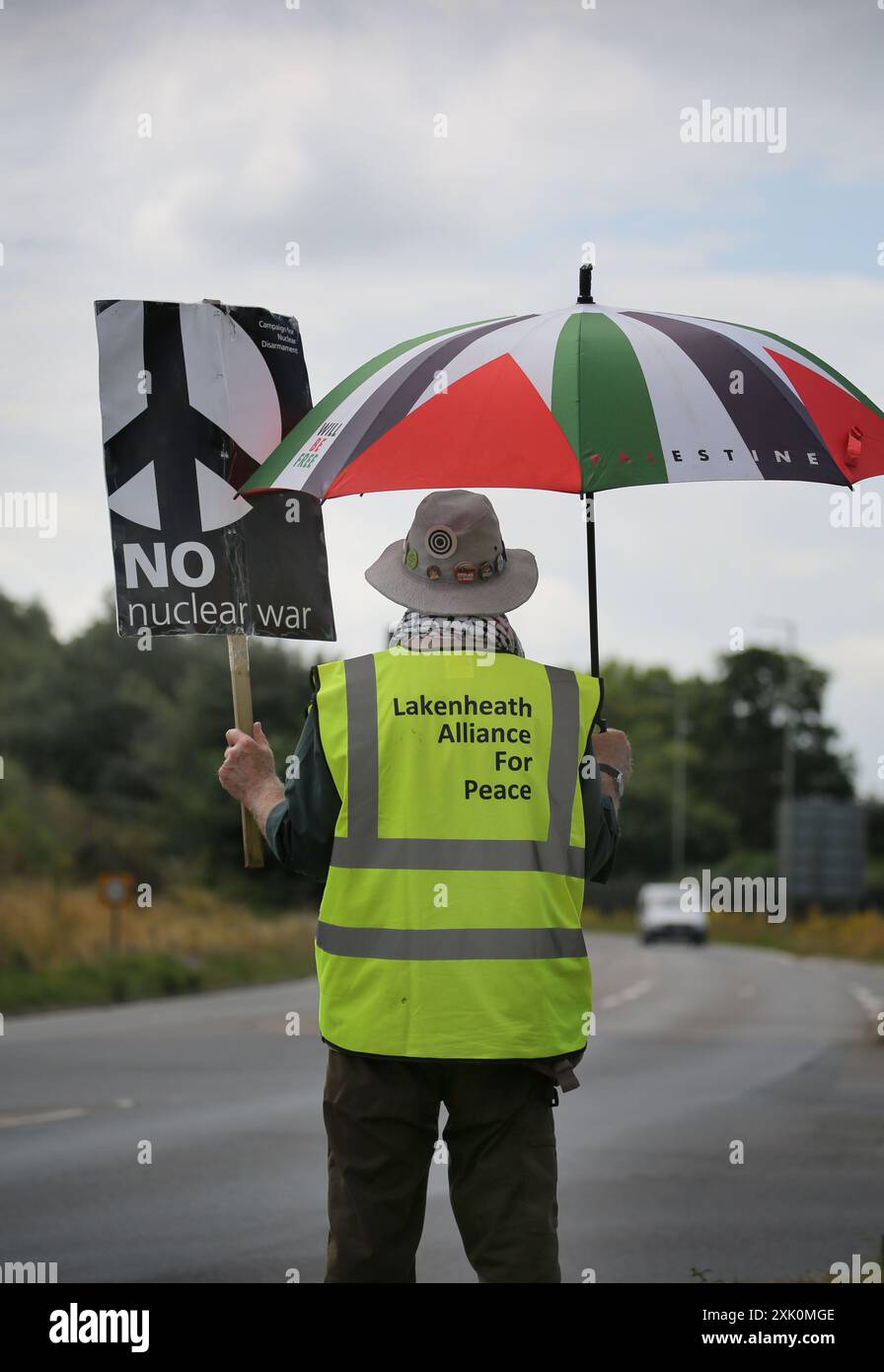 Lakenheath, England, Großbritannien. Juli 2024. Ein Demonstrant steht neben der Hauptstraße vor dem Luftwaffenstützpunkt in Lakenheath. Mit einem Schild mit der Aufschrift "kein Atomkrieg" und einem palästinensischen Unterstützungsschirm während der Demonstration. Der Protest wurde von der Campaign for Nuclear Disarmament (CND) organisiert, deren Unterstützer sich gegen die mögliche Rückgabe von Atomwaffen auf Militärbasen in Großbritannien, wie etwa die RAF Lakenheath, aussprechen, nachdem berichtet wurde, dass die USA beabsichtigen, Atombomben auf der Basis zu widersetzen. Nach anhaltenden Protesten wurden 2008 Atomwaffen aus Lakenheath entfernt. (Bild: © Martin Pop Stockfoto