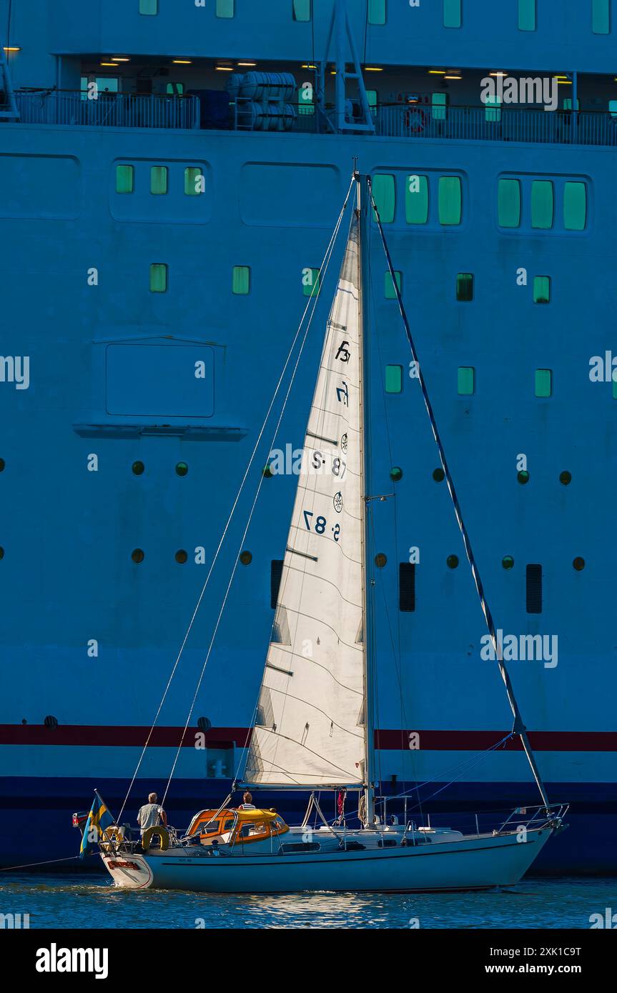 Ein kleines Segelboot mit weißem Segel navigiert durch das Wasser vor dem großen Schiff Göteborg. Das Boot ist von zwei Personen besetzt. Die Szene ist baden Stockfoto