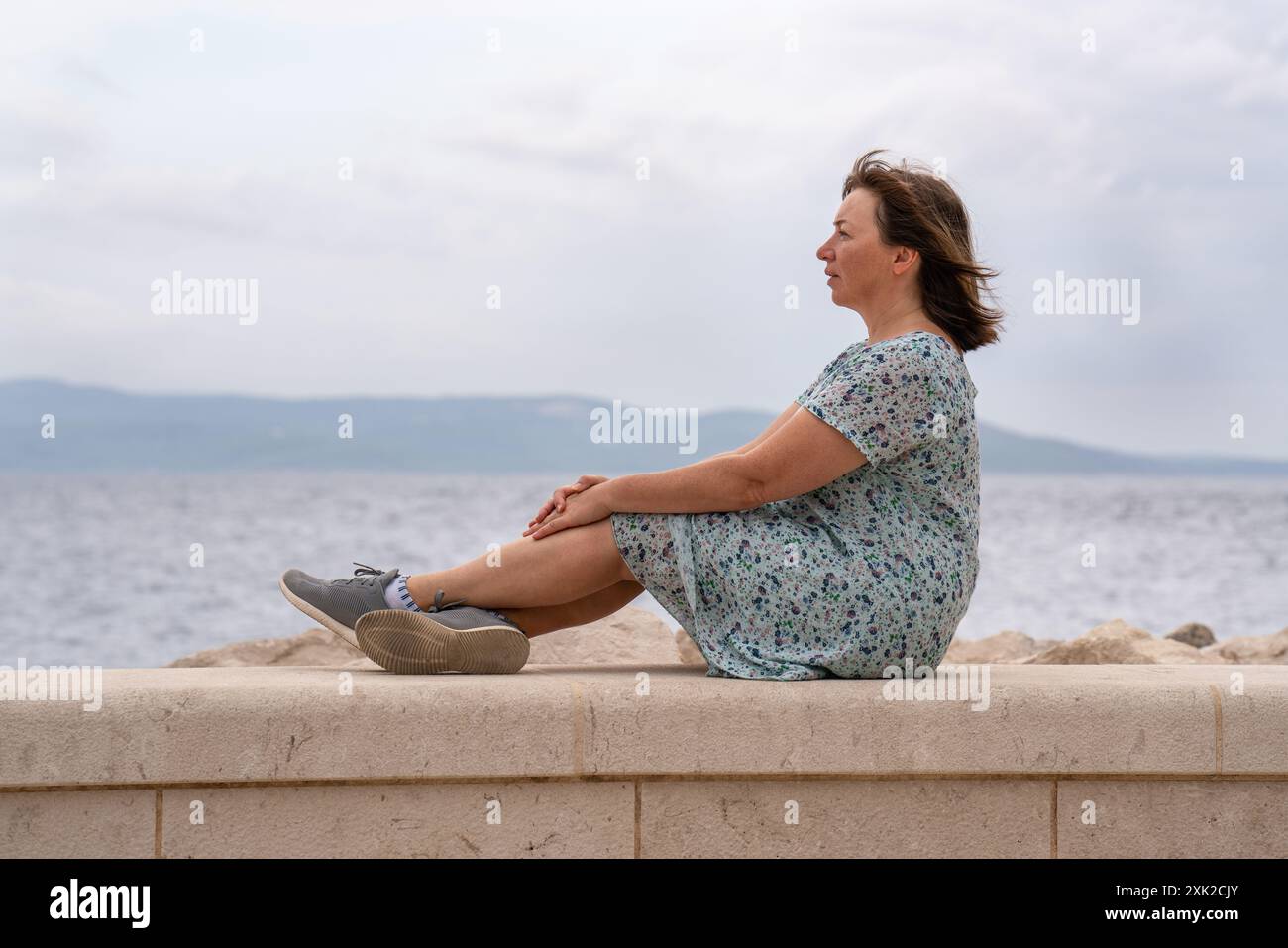 Eine Frau mittleren Alters sitzt am Meer, auf dem Pier und blickt in die Ferne. Einsamkeit, Nachdenklichkeit, Einsamkeit. Meer und Berge auf den Hori Stockfoto