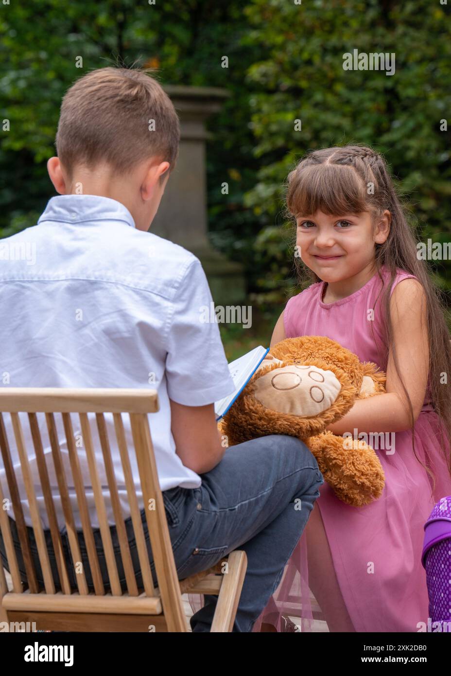 Der Junge sitzt mit dem Rücken auf einem Stuhl und liest ein Buch. Das Mädchen schaut mit Interesse rein und will sich die Bilder ansehen. Mädchen, 6 Jahre alt, mit einer Stockfoto