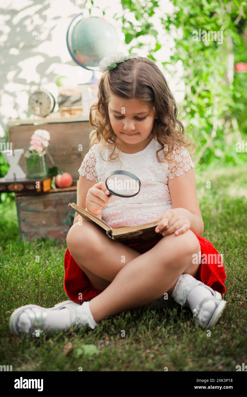 Ein junges Mädchen sitzt im Kreuz auf dem Gras und liest mit einer Lupe ein Buch. Sie trägt ein weißes Hemd und einen roten Rock. Back to School Conce Stockfoto