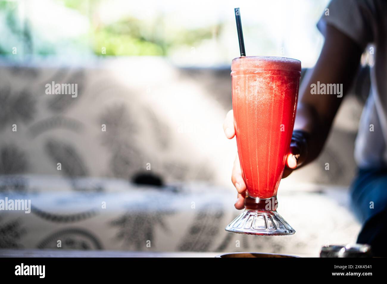 Eine Person, die ein hohes Glas rotes Getränk mit einem Strohhalm hält, vor einem verschwommenen Hintergrund eines gemütlichen Cafés. Das Getränk wirkt erfrischend und schaumig, Stockfoto