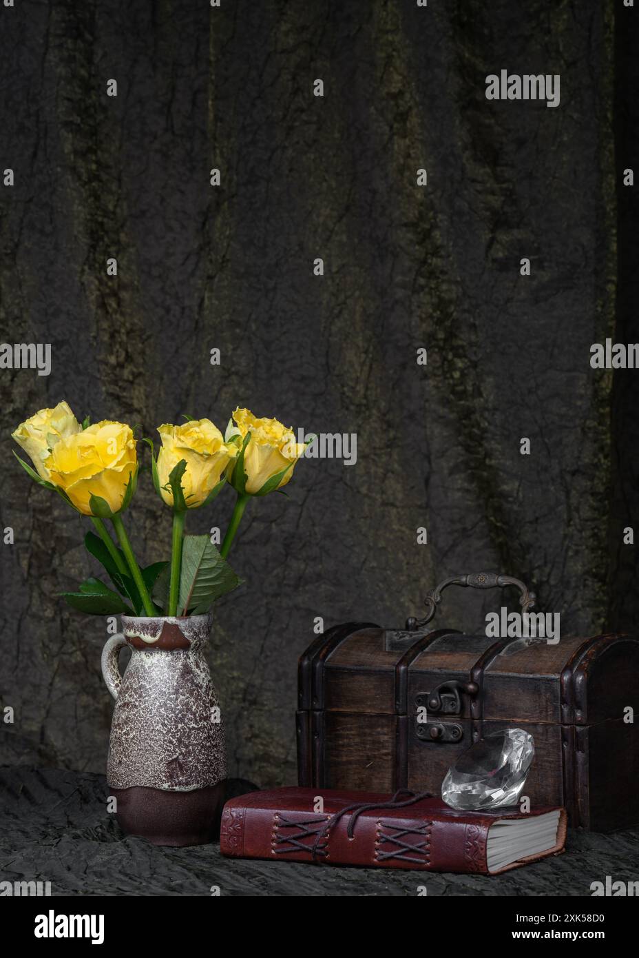 Gelbe Rosen in einer Vase vor dunklem Stoff, neben einer alten Holzkiste. Buch und Juwel mit Lederbindung. Platz für Text. Rätsel, Rätsel Stockfoto
