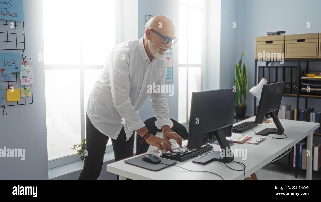 Mann, der an einem Computer in einem modernen Büro mit Bart und Brille arbeitet, umgeben von organisierten Regalen und motivierenden Zitaten. Stockfoto
