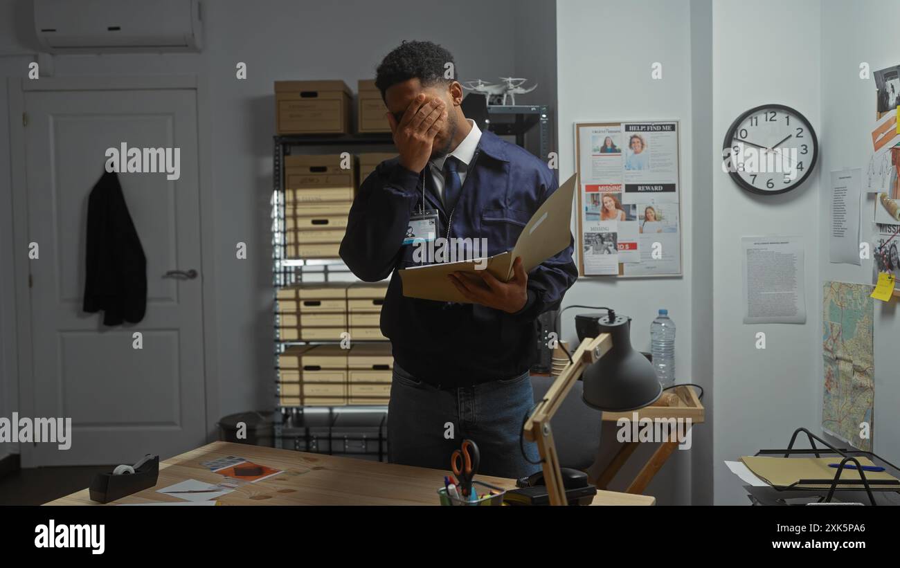 Ein afroamerikanischer Detective erscheint gestresst in einem überfüllten Polizeibüro und hält eine Akte mit Beweismitteln neben einer Tafel. Stockfoto