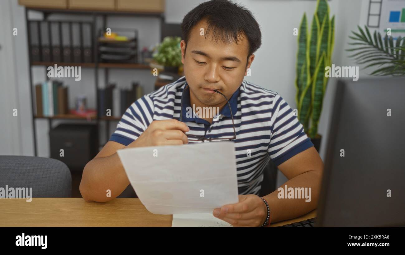 Hübscher junger chinesischer Mann, der in einem Büro arbeitet, liest ein Dokument aufmerksam, während er seine Brille hält, asiatischer Erwachsener, der sich an einem Innenarbeitsplatz konzentriert Stockfoto