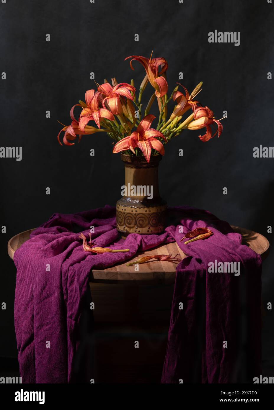 Ein Strauß orangener Lilien in einer grünen Vase, neben einem lila Schal. Grauer Hintergrund. Stillleben Stockfoto