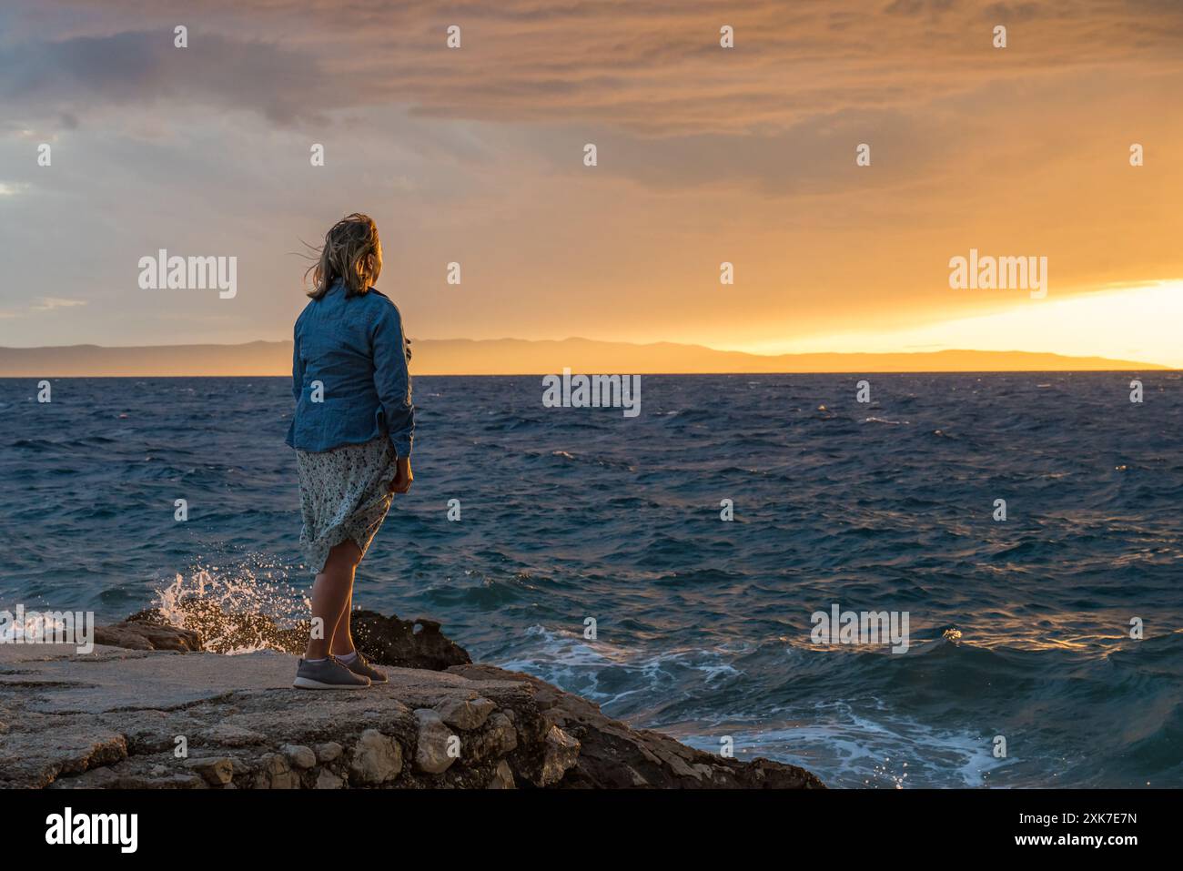 Eine Frau mittleren Alters, die das Leben genießt. Am Rande des Strandes stehen und den wunderschönen gelb-orangen Sonnenuntergang auf dem Meer betrachten. Wellen, die auf t abstürzen Stockfoto