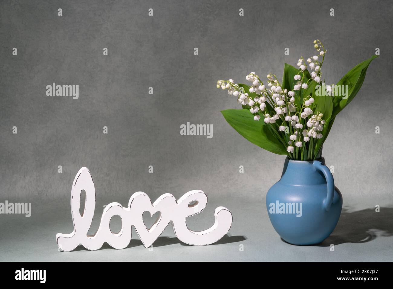 Ein kleiner Strauß weißer Blumen, Maiglöckchen aus dem Tal in einer blauen Vase neben der Aufschrift „Liebe“. Grauer Hintergrund. Platz für Text. Stillleben. Pos. Stockfoto