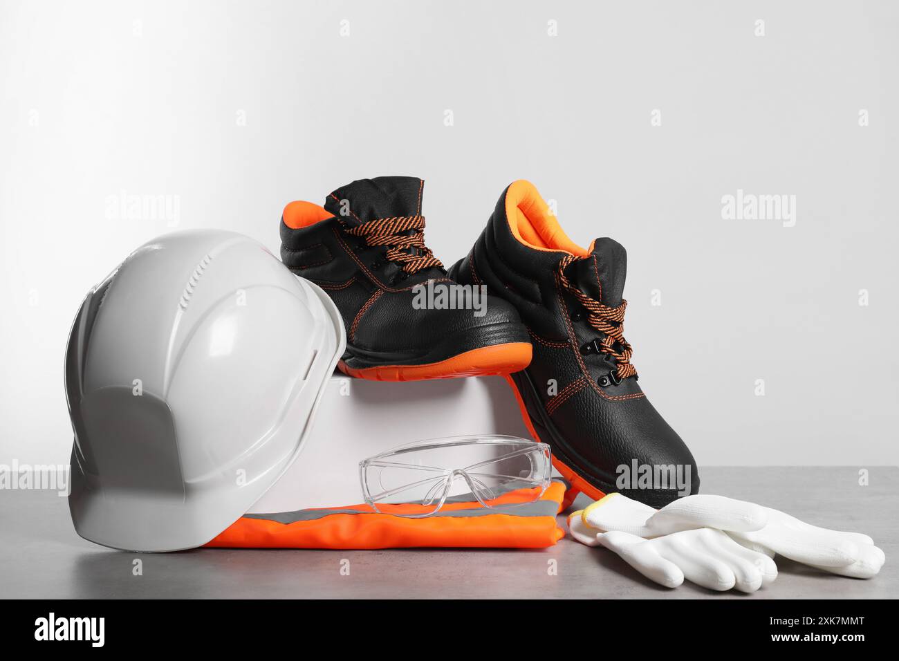 Schutzhelm, Paar Arbeitsstiefel, Schutzhandschuhe, Schutzbrille und reflektierende Weste auf grauer Oberfläche vor hellem Hintergrund Stockfoto