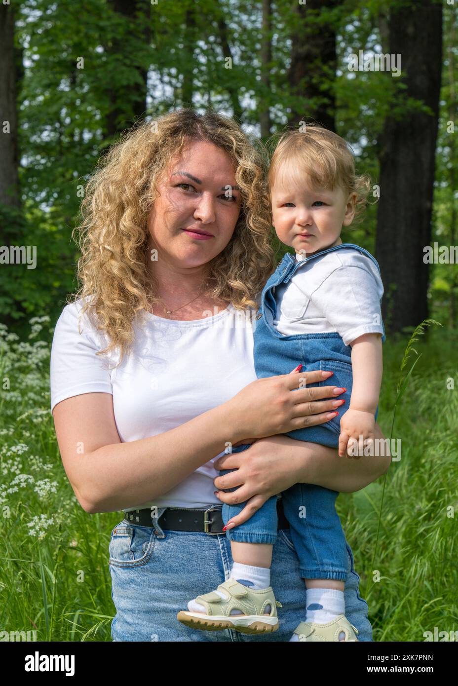 Porträt von Mutter und Sohn. Eine glückliche Mutter mit welligen Haaren liegt auf dem grünen Gras zwischen weißen Gänseblümchen, ein kleines Baby mit welligen, blonden Haaren sitzt Stockfoto