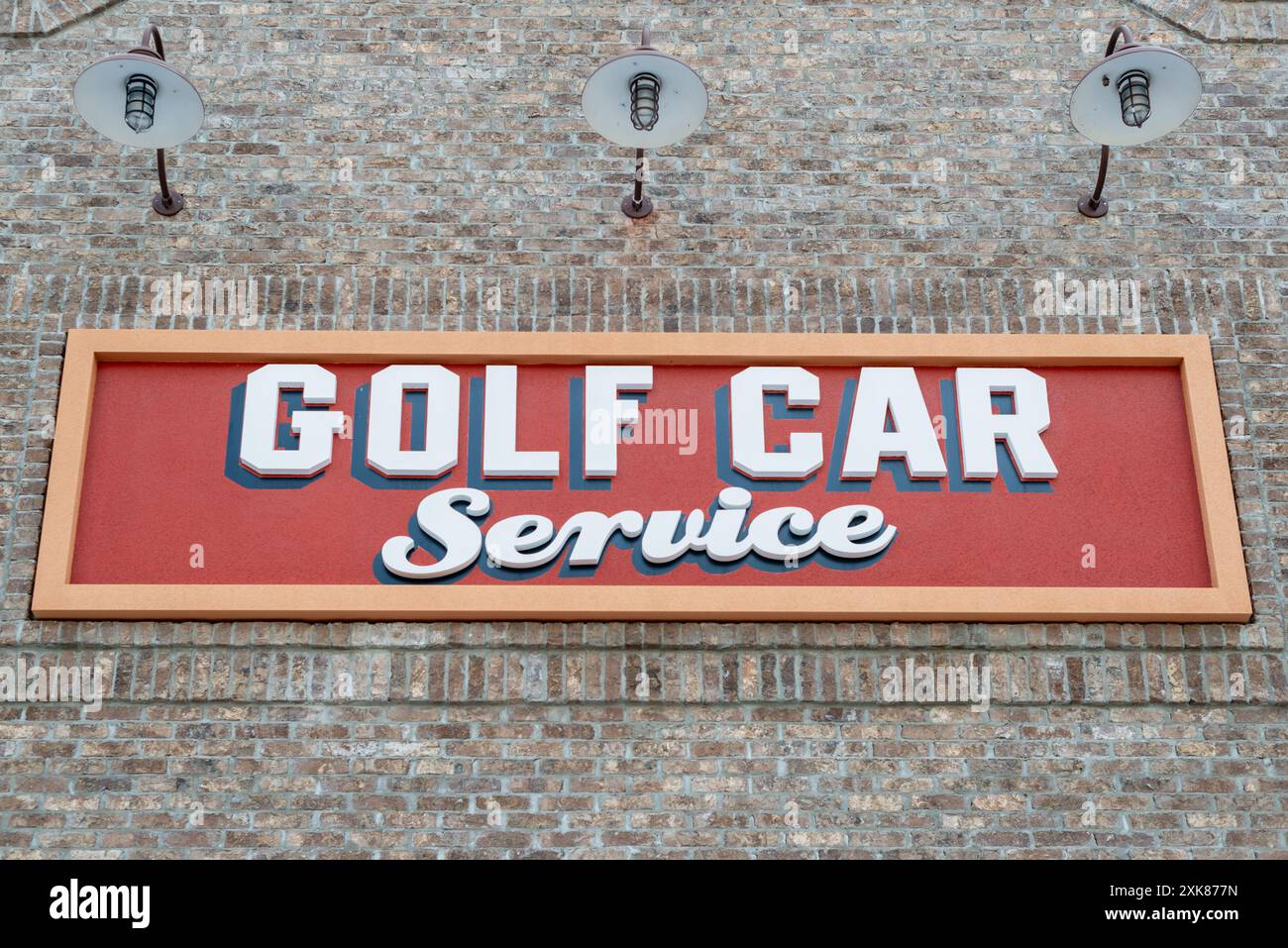 Ein rot-gelbes Golf Car Service Schild mit weißem Text und schwarzer Zierleiste. Das Schild ist an einem braun-beigefarbenen Ziegelgebäude mit drei Scheunen angebracht Stockfoto