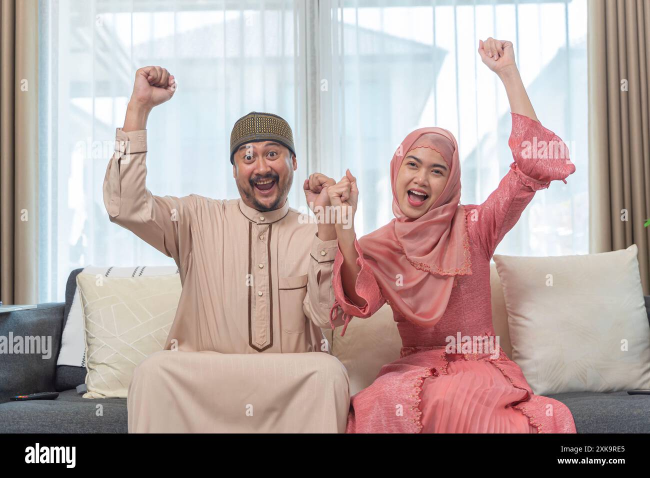 Glückliches arabisches Paar in islamischer Kleidung, das auf dem Sofa im Wohnzimmer sitzt und Freude zeigt mit erhobenen Händen und lächelnden Gesichtern, glückliches Paar erhält gute Nachrichten. Stockfoto