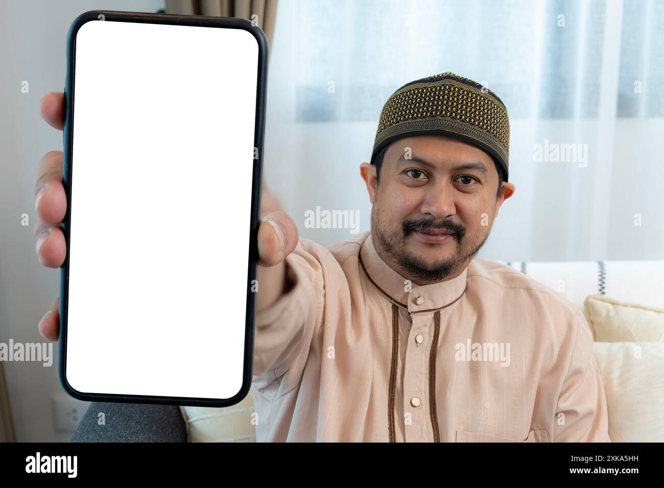 Junger arabischer Kerl, der neben einem großen Handy sitzt, mit leerem weißen Bildschirm, mit Mobilgerät, Mockup. Lächelnder junger arabischer Mann, der ein riesiges Smartphone demonstriert Stockfoto