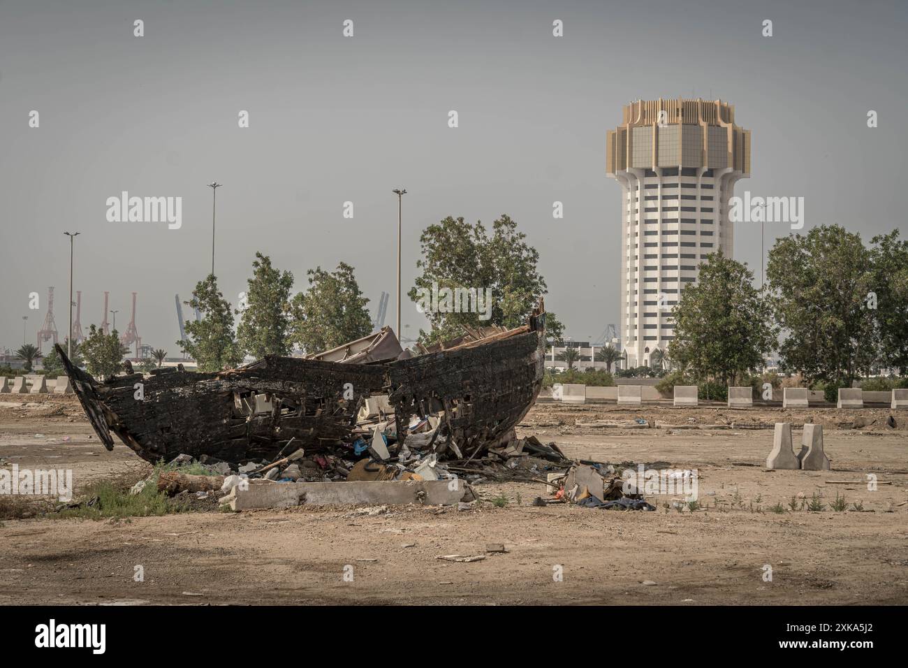 Das alte verlassene Boot an der Trockenküste von Dschidda, Saudi-Arabien, mit dem Gebäude des islamischen Hafens von Dschidda im Hintergrund. Stockfoto