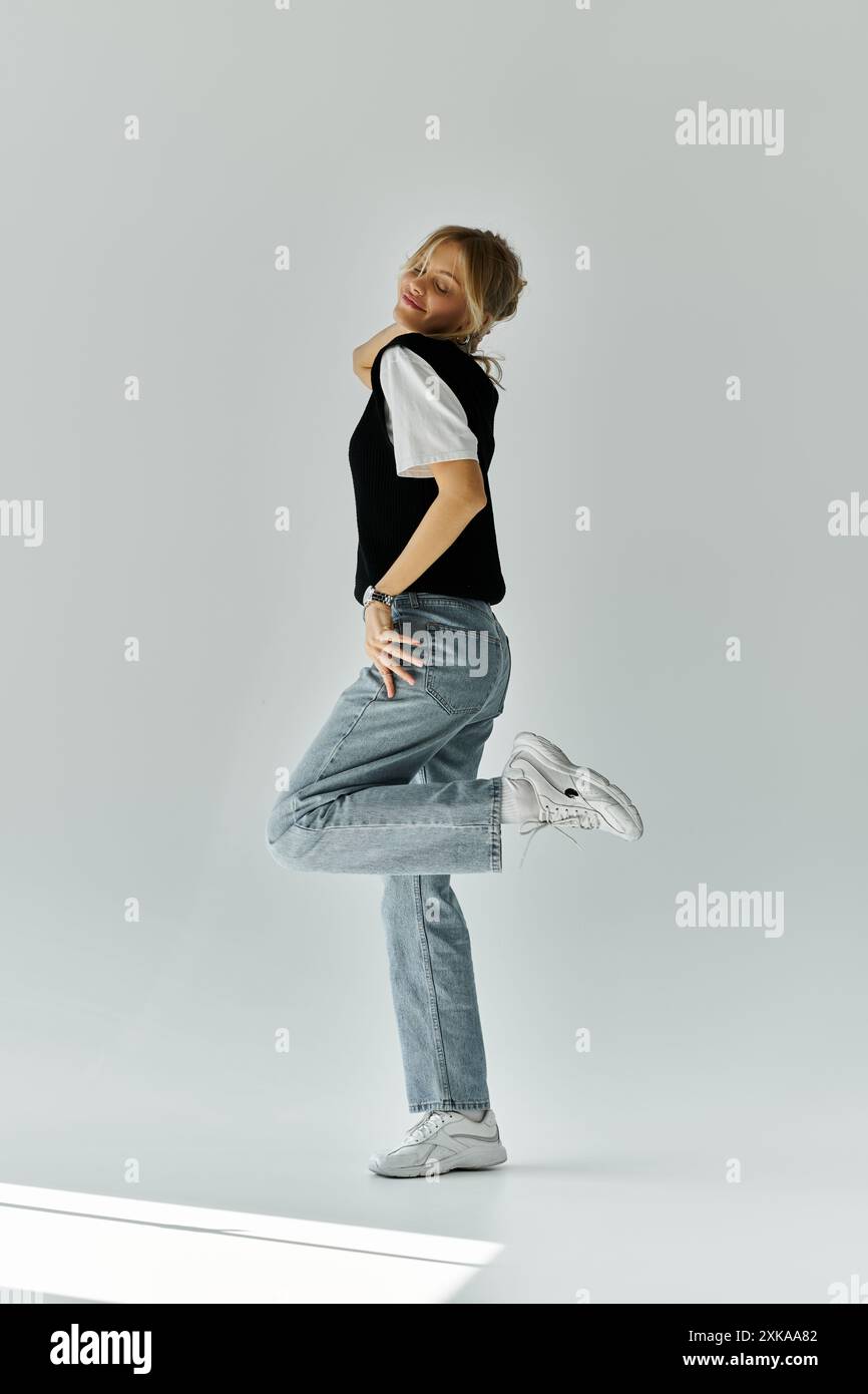 Eine junge Frau mit blonden Haaren posiert in einem schwarzen Hemd und einer Jeans vor grauem Hintergrund. Stockfoto