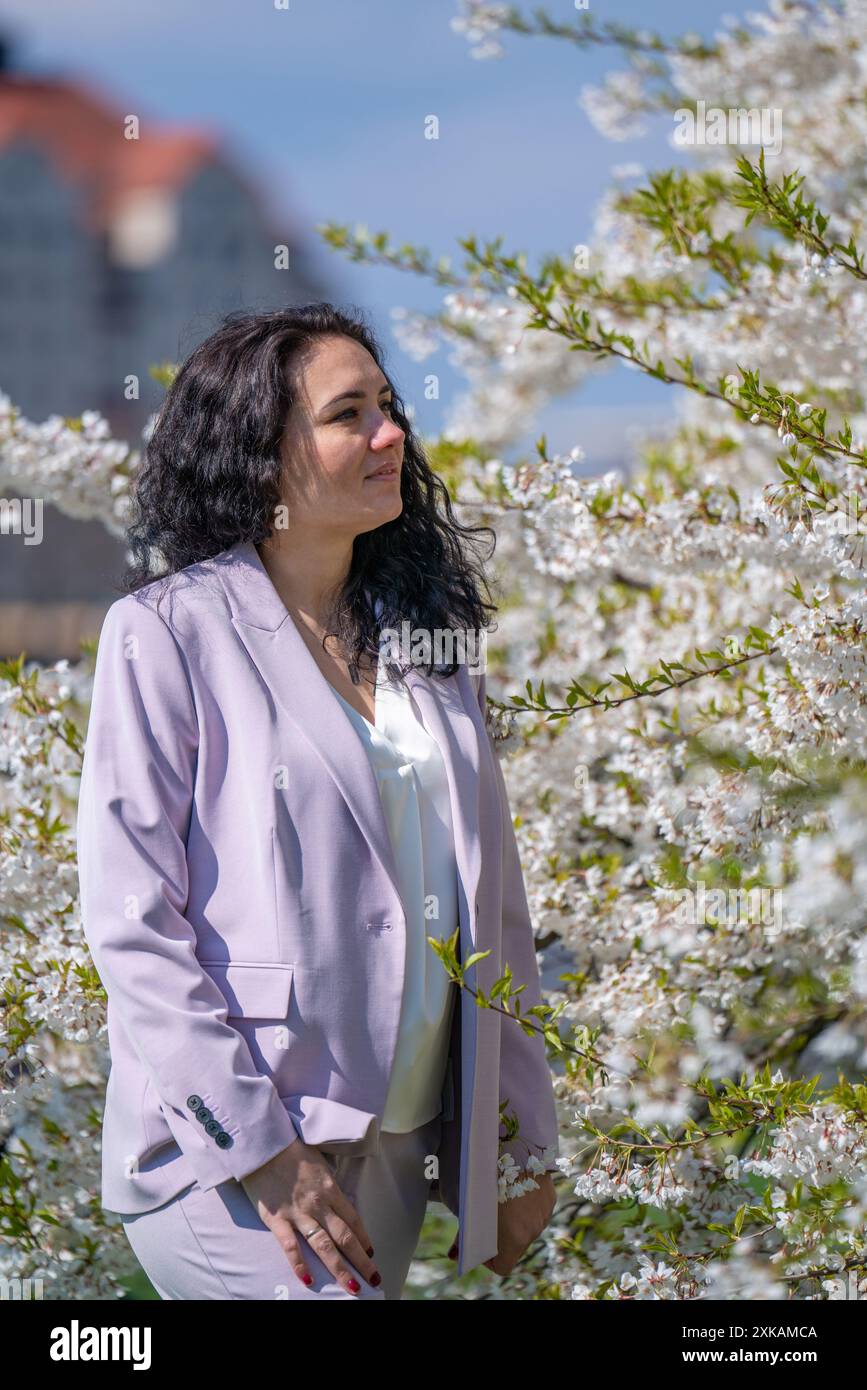 Romantisches Bild einer stilvollen Frau in einer leichten Jacke. Positive Frühlingsstimmung. Ein süßes Mädchen hält sanft einen weißen Sakura-Zweig und schaut sich die Blumen an Stockfoto