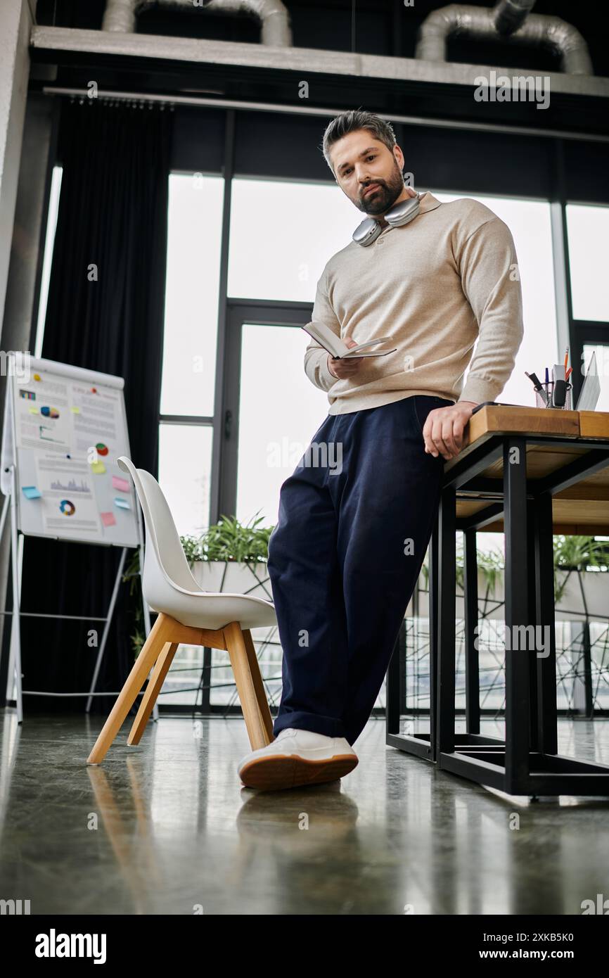 Ein gutaussehender Geschäftsmann mit Bart lehnt sich in einem modernen Büro lässig an einen Schreibtisch und überprüft ein Dokument sorgfältig. Stockfoto