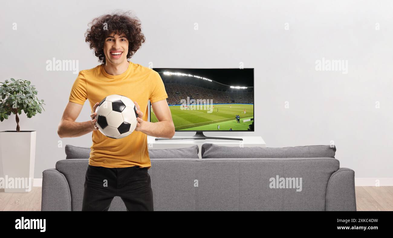 Junger leidenschaftlicher Fußballfan, der einen Ball hält und zu Hause vor dem fernseher in die Kamera lächelt Stockfoto