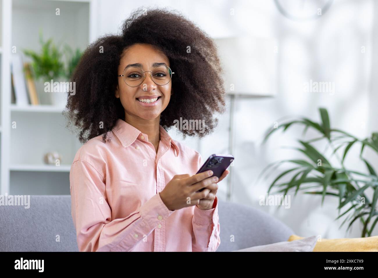 Glückliche junge Frau mit lockigem Haar und Brille, die ihr Smartphone benutzt, während sie im gemütlichen Wohnzimmer auf der Couch sitzt. Ungezwungene Indoor-Szene mit Person, die wi Stockfoto Glückliche junge Frau mit lockigem Haar und Brille, die ihr Smartphone benutzt, während sie im gemütlichen Wohnzimmer auf der Couch sitzt. Ungezwungene Indoor-Szene mit Person, die wi Stockfoto