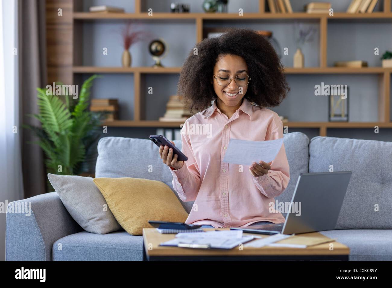 Lächelnde Frau, die mit Smartphone und Laptop auf dem Sofa sitzt und gleichzeitig Haushaltsfinanzen verwaltet. Entspanntes Wohnumfeld mit positiver finanzieller Stimmung Stockfoto