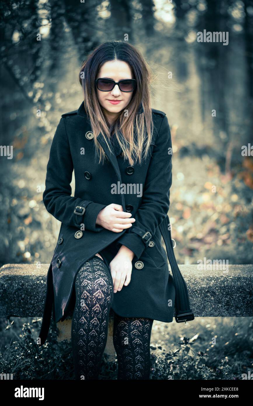 Eine junge Frau mit schwarzem Mantel und oberschenkelhohen Strümpfen sitzt auf einem verwitterten Baumstamm in einer Waldlandschaft. Ihr dunkles Haar umrahmt ihr Gesicht und sie trägt Stockfoto