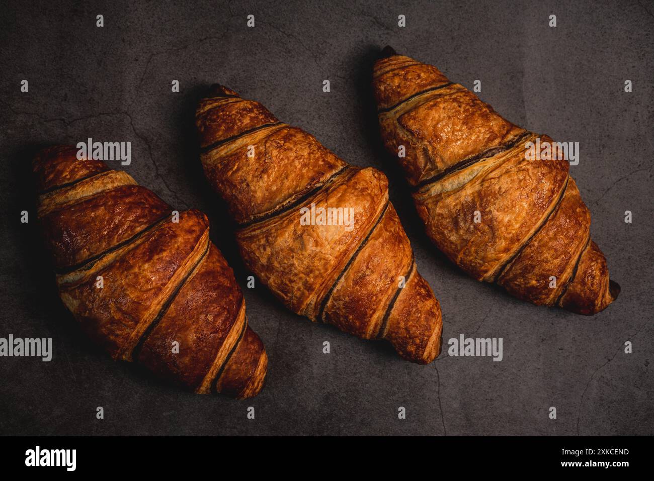 Drei frisch gebackene Croissants sitzen auf einer dunkelgrauen Arbeitsplatte, bereit zum Verzehr. Ihre goldbraune Kruste glitzert unter dem weichen Licht und lädt ein Stockfoto