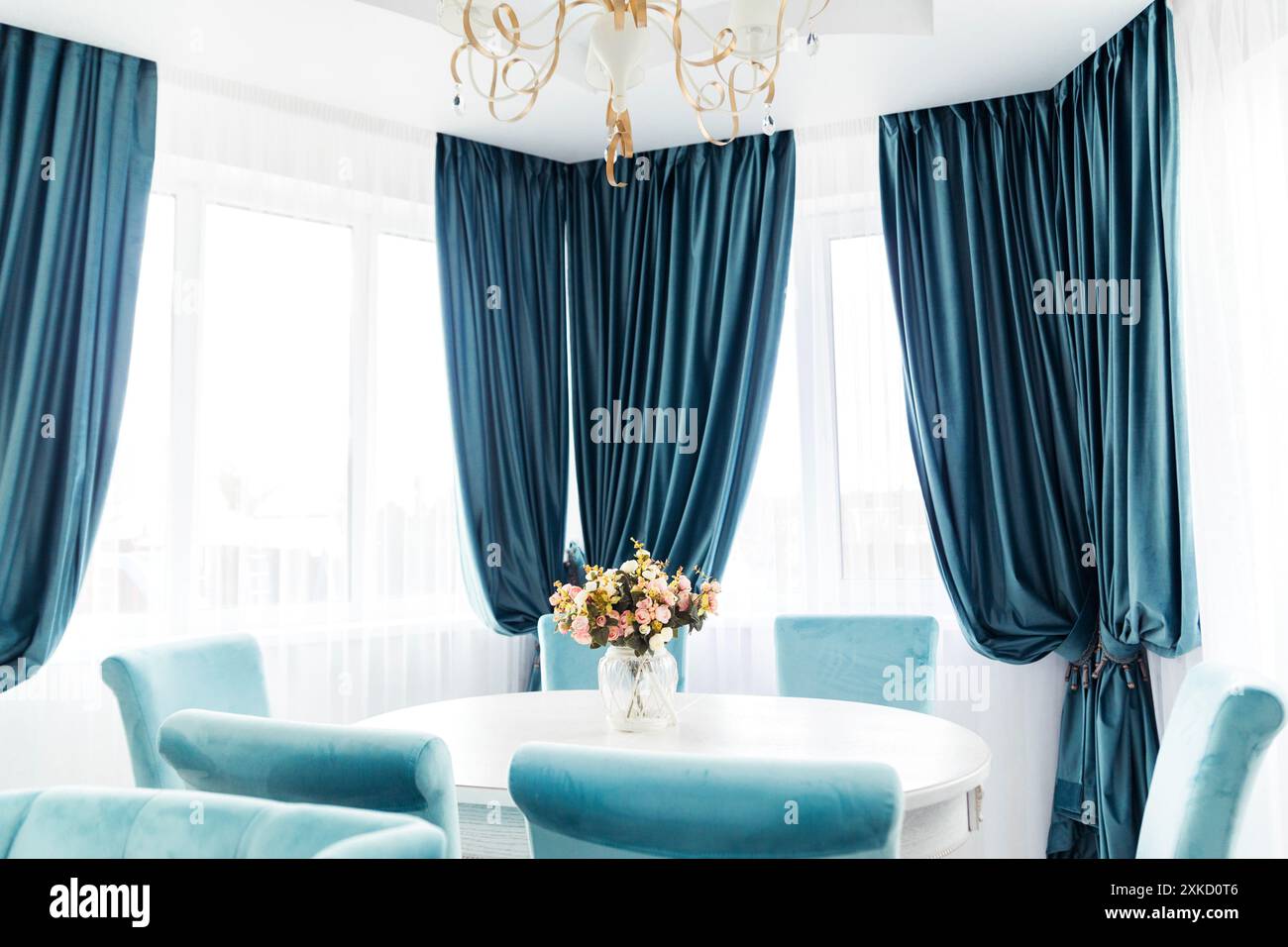 Heller Speisesaal mit blauen Samtvorhängen, passenden Stühlen und einem weißen Tisch. Elegantes und luftiges Innendesign. Stockfoto