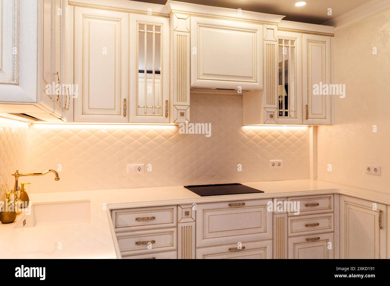 Elegante weiße Küche mit goldenen Akzenten, maßgefertigten Schränken, modernen Geräten und gefliestem Backsplash. Die warme Beleuchtung sorgt für ein raffiniertes Ambiente Stockfoto