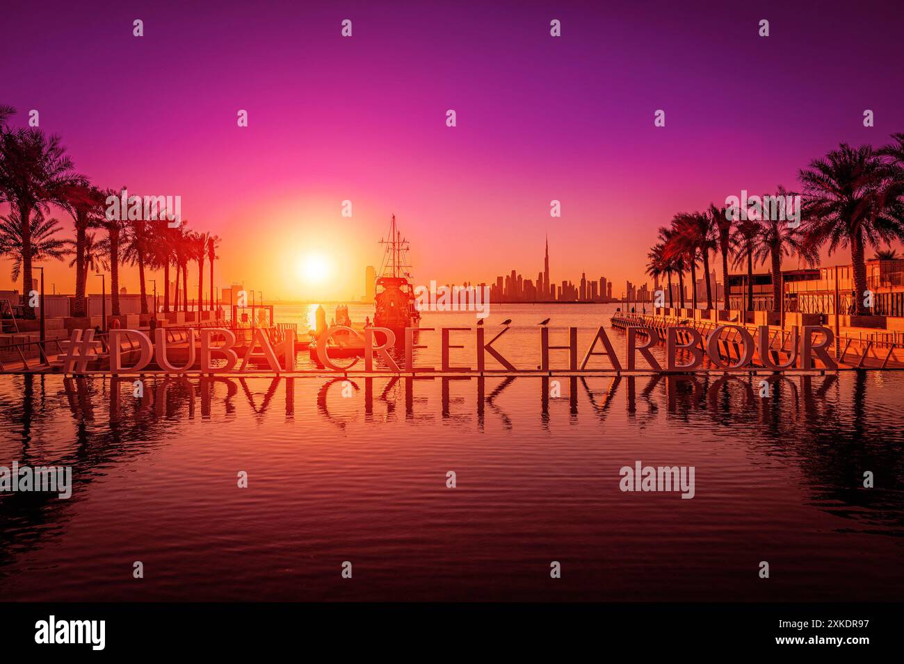 Ein großes Schild für den Hafen von Dubai Creek befindet sich bei Sonnenuntergang im Wasser, mit Palmen und einer Skyline der Stadt im Hintergrund. Stockfoto