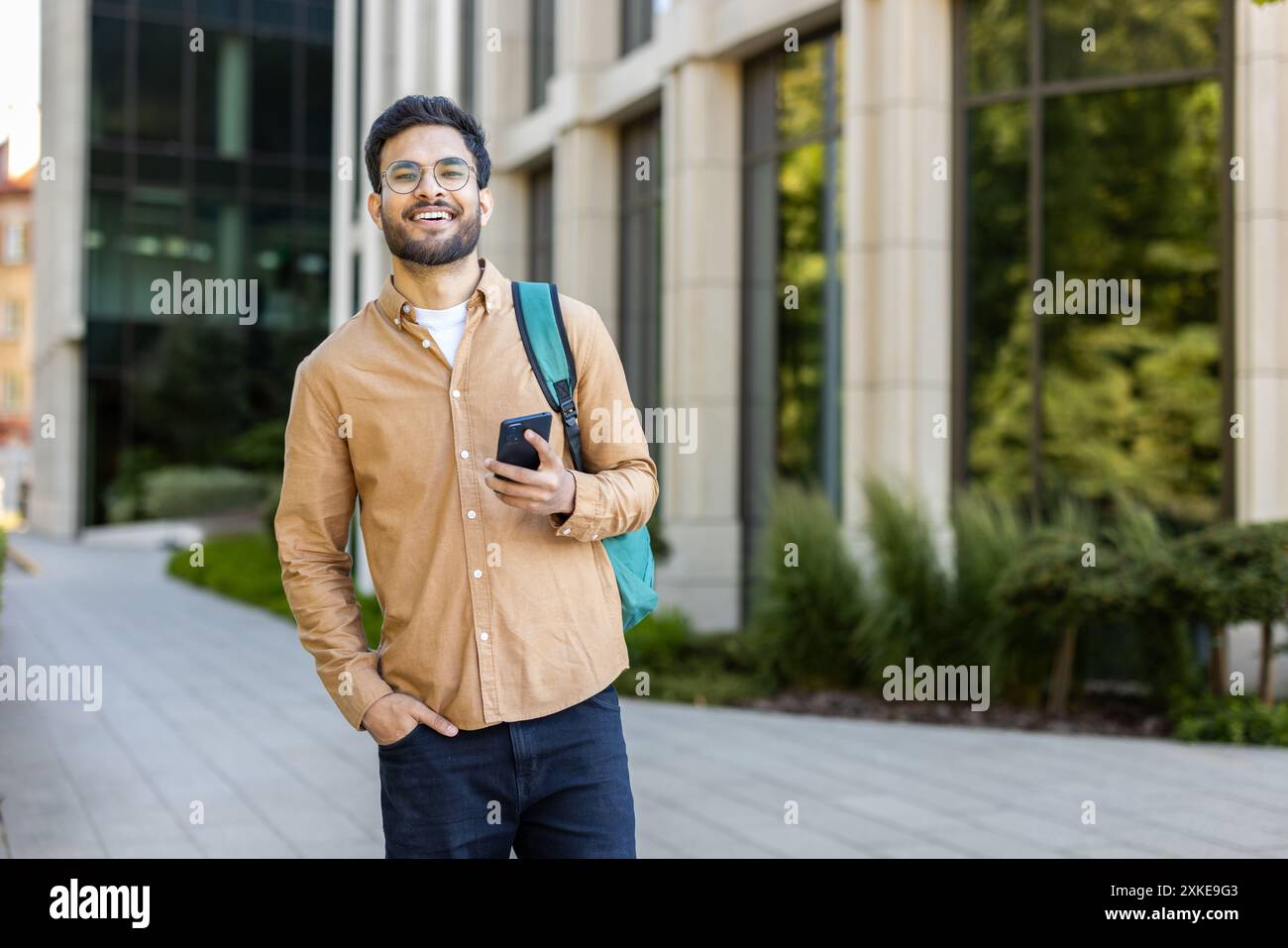 Selbstbewusster junger indischer Student, der ein Smartphone hält, lächelt und draußen auf einem sonnigen Universitätscampus steht. Das Konzept der Bildung, Jugend und Technologie. Stockfoto