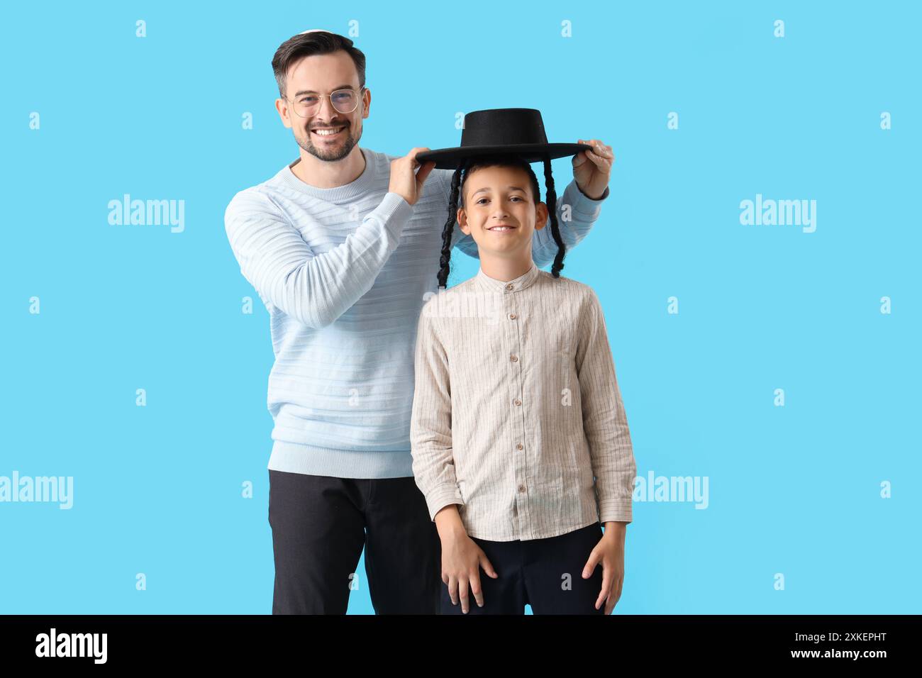 Glücklicher jüdischer Vater, der seinen kleinen Sohn vor blauem Hintergrund mit Hut bedeckt Stockfoto