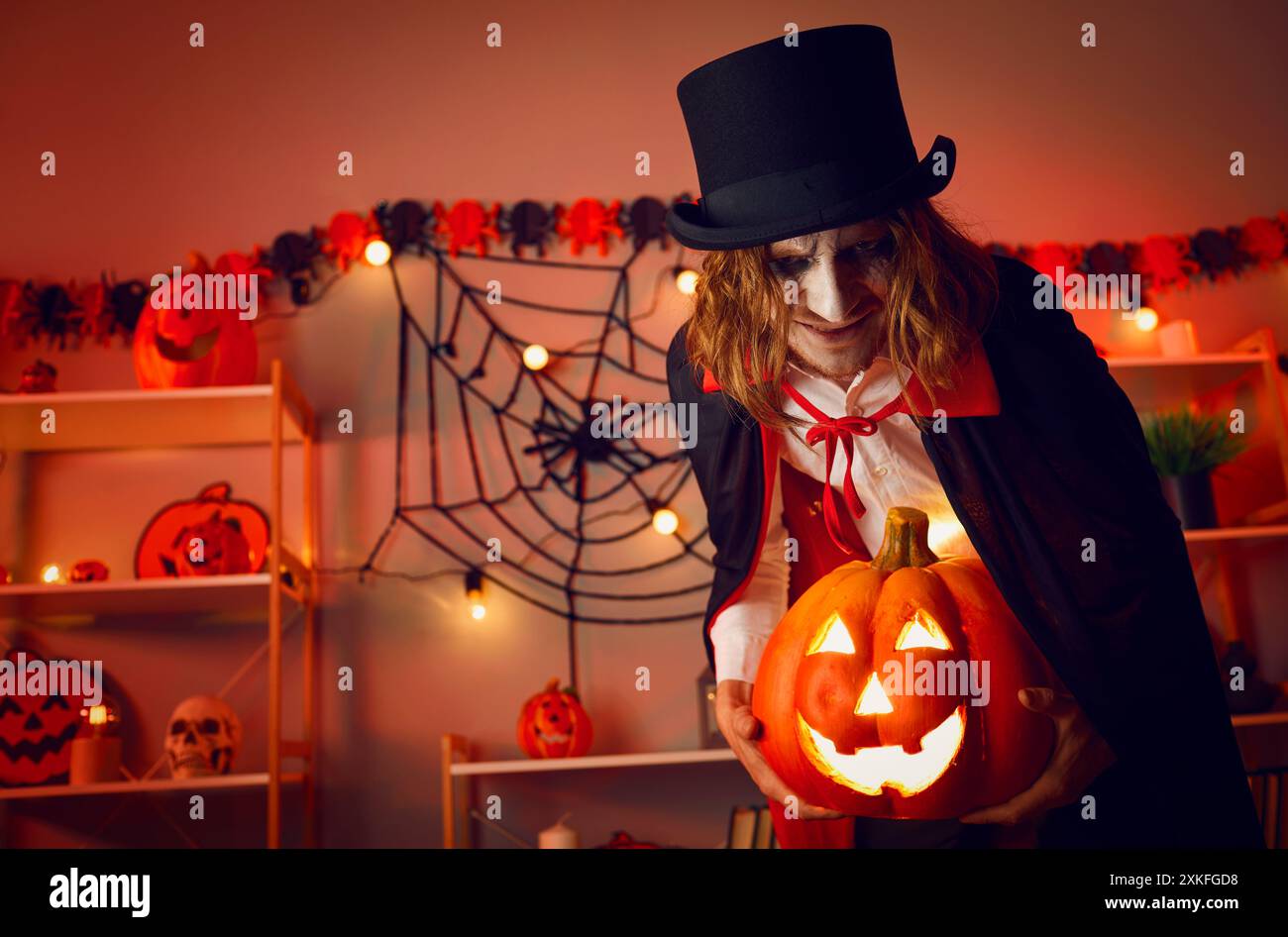 Gruseliges Porträt eines Vampirmannes, der im Dunkeln im Zimmer steht, mit leuchtender Jack-o-Laterne in den Händen. Stockfoto