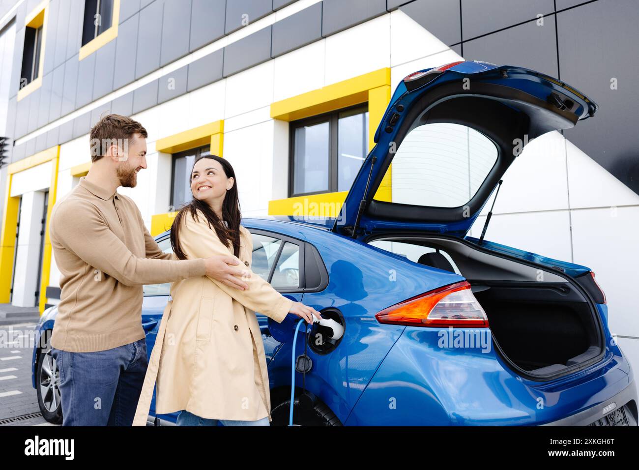 Ein junges Paar, das mit einem Elektroauto unterwegs ist und an der Ladestation hält. Eine Freundin, die das Ladegerät hält, das Auto ansieht und lächelt. Stockfoto