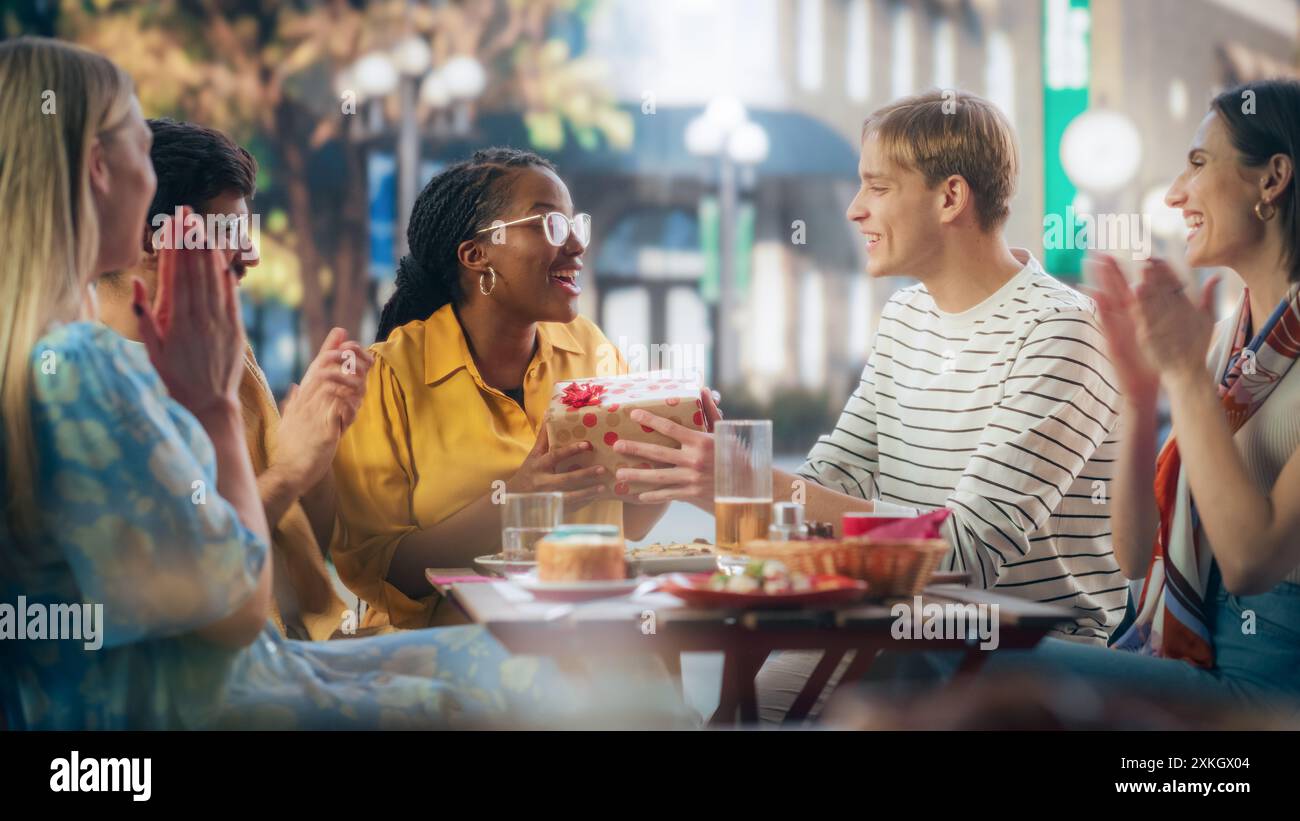 Schöne Freunde versammelten sich, um ein Geburtstagsfest in einem Street Cafe zu feiern. Eine Gruppe junger Frauen und Männer überreicht ein Geschenk an ein glückliches schwarzafrikanisches Mädchen. Freunde, die eine Party auf einer Terrasse feiern Stockfoto