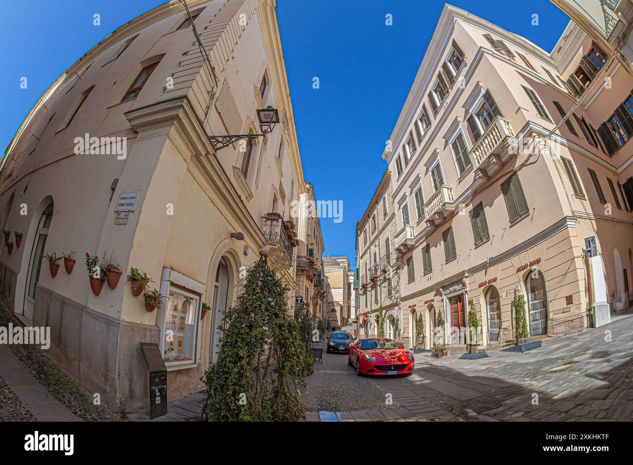 ALGHERO, ITALIEN - 4. JULI 2024: Moderne Boliden auf der schmalen typisch sardischen Straße, Piazza Civica, mit dem Gebäude des Palazzo Bolasco an der Kreuzung Stockfoto