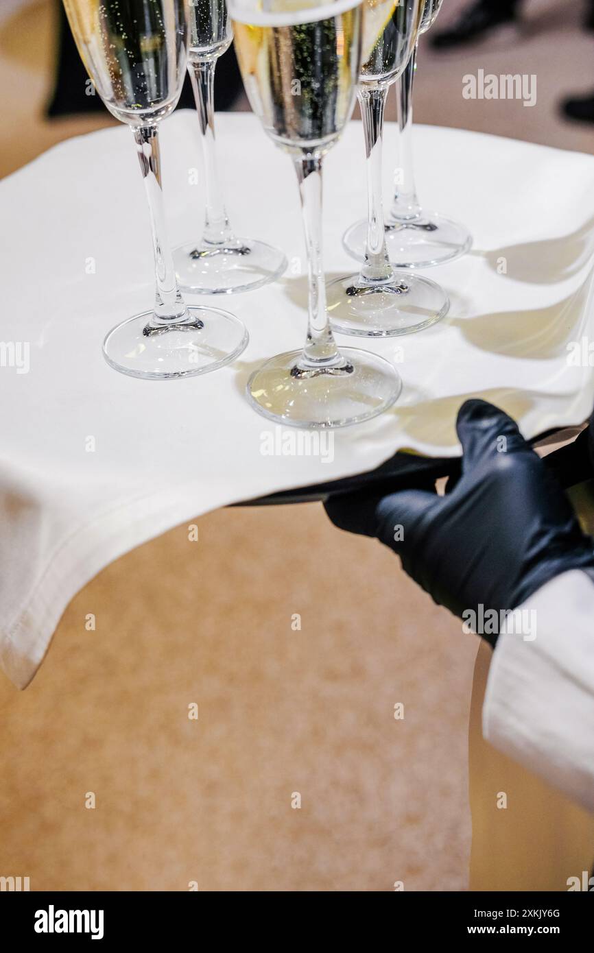 Nahaufnahme einer Hand mit Handschuhen, die ein Tablett mit Sektflöten hält, das für Gäste bei einer formellen Veranstaltung bereit ist. Vertikales Foto Stockfoto