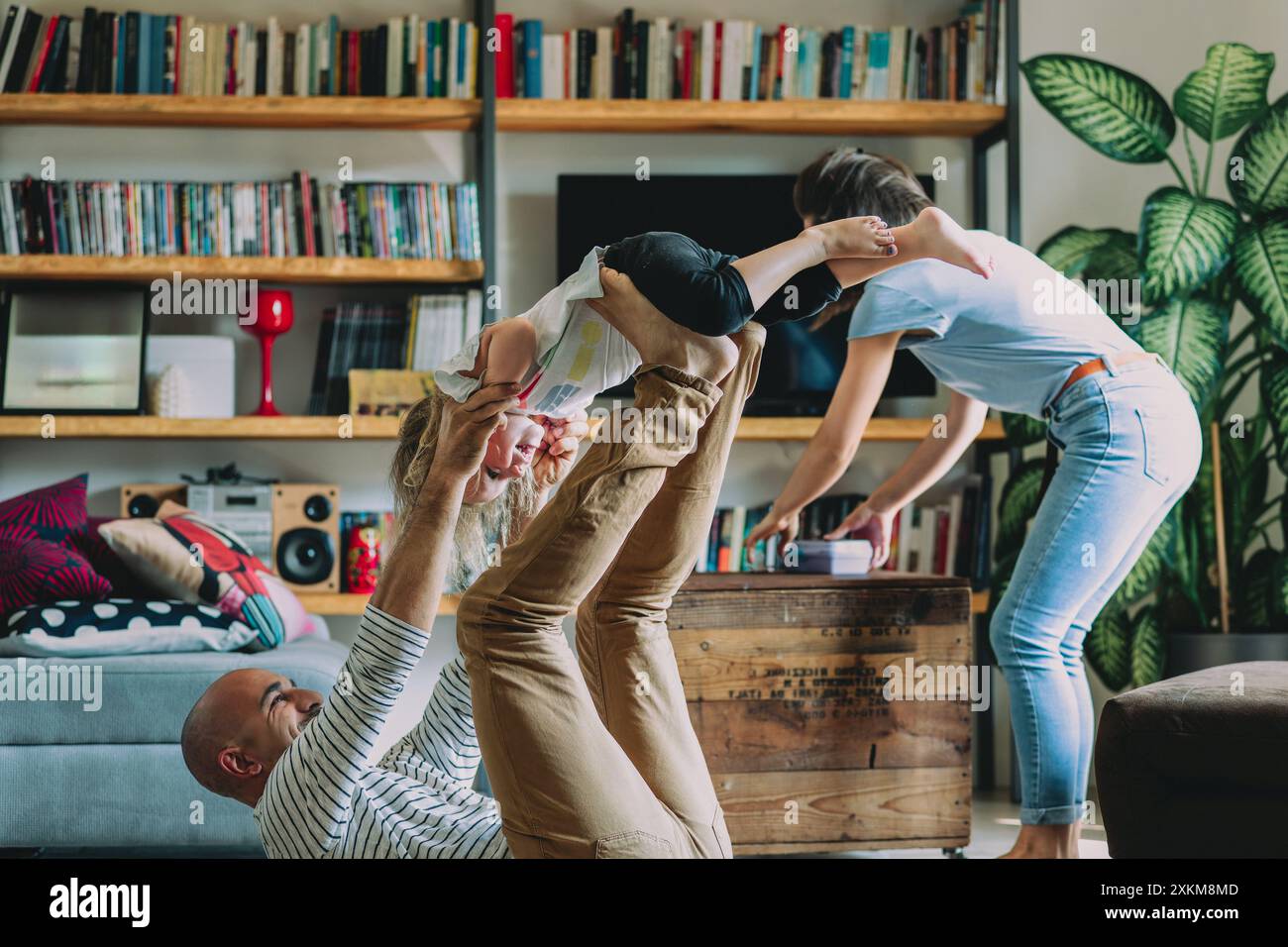Fröhliche Familie, die in einem gemütlichen Wohnzimmer mit Möbeln und Pflanzen spielt und sich zusammensetzt und freudige Erinnerungen schafft Stockfoto