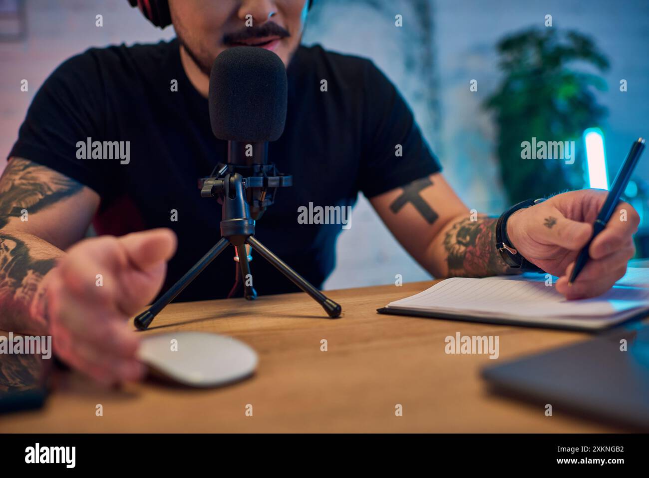 Ein asiatischer Mann in einem Studio, der einen Podcast aufnimmt, mit einem Mikrofon vor ihm und einem Notizbuch und Stift neben ihm. Stockfoto