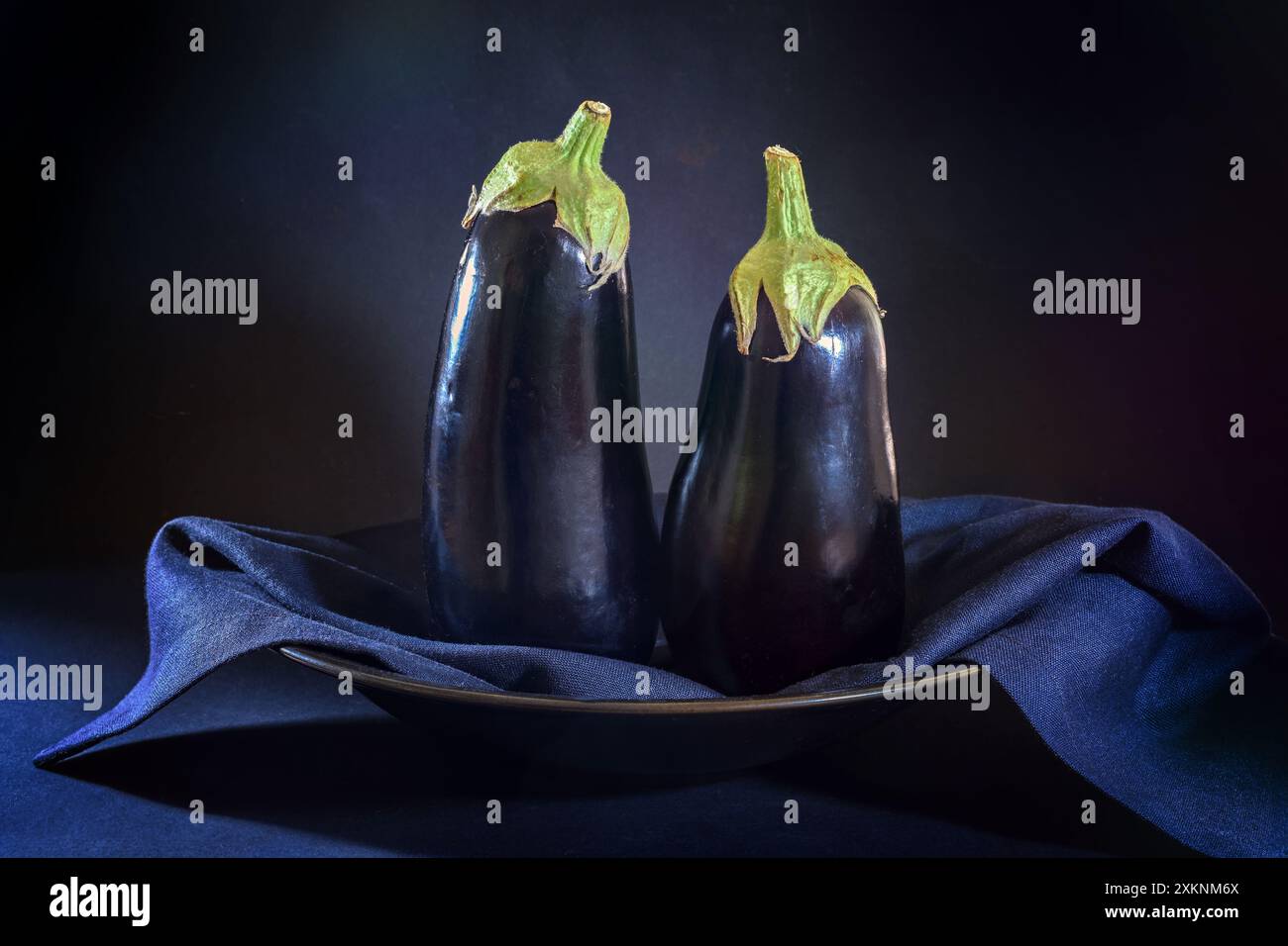 Zwei Auberginen stehen auf einem Teller mit einer dunkelblauen Serviette vor fast schwarzem Hintergrund, gesundes Gemüse wie eine Kunstskulptur angeordnet, Kopie sp Stockfoto