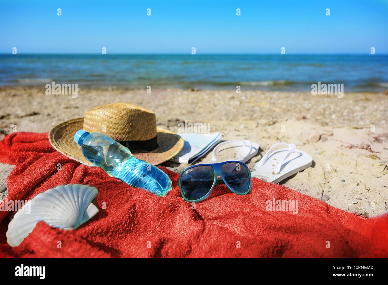 Badeurlaub am Strand mit rotem Handtuch, Wasserflasche, Strohhut, Sonnenbrille und Flipflops, Ostsee in Deutschland, Kopierraum, ausgewählter Fokus Stockfoto