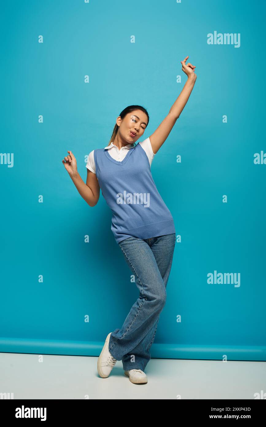 Eine junge asiatische Frau in lässiger Kleidung posiert vor einem hellblauen Hintergrund, ihre sorglose Haltung strahlt Glück aus. Stockfoto