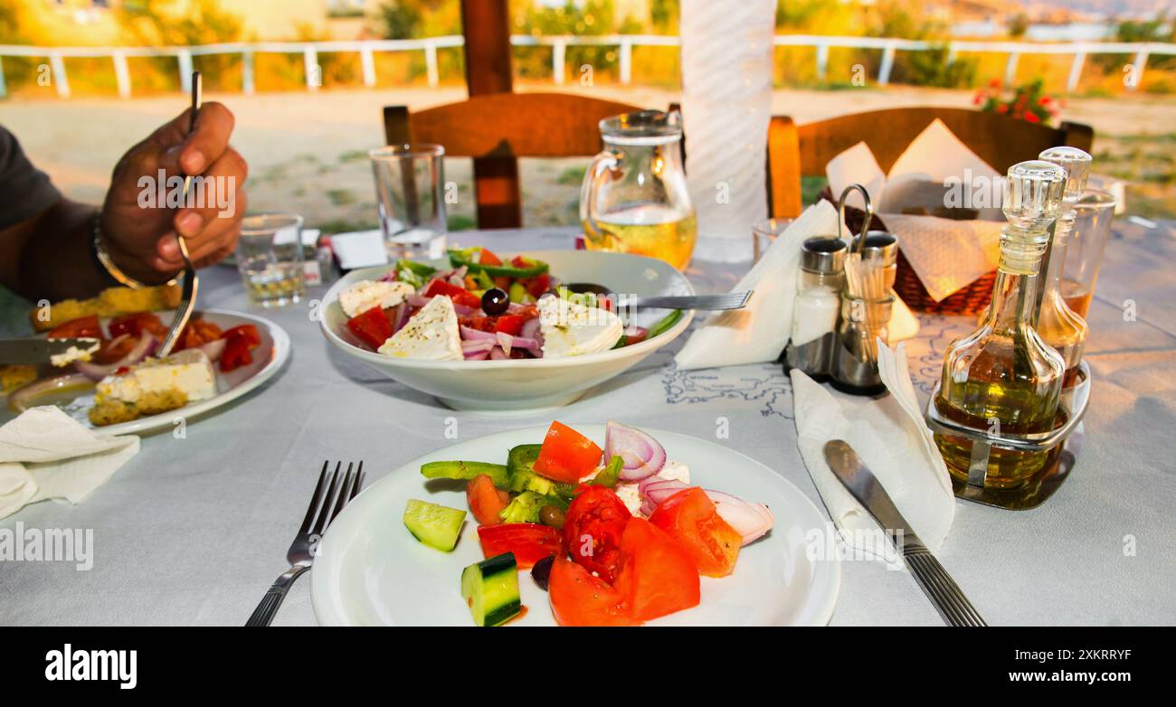 Speisen im griechischen Restaurant im Freien, Teller mit frischem griechischen Salat, Olivenöl, Schüssel. Ein Teil der Hand des Mannes, isst er Salat. Stockfoto