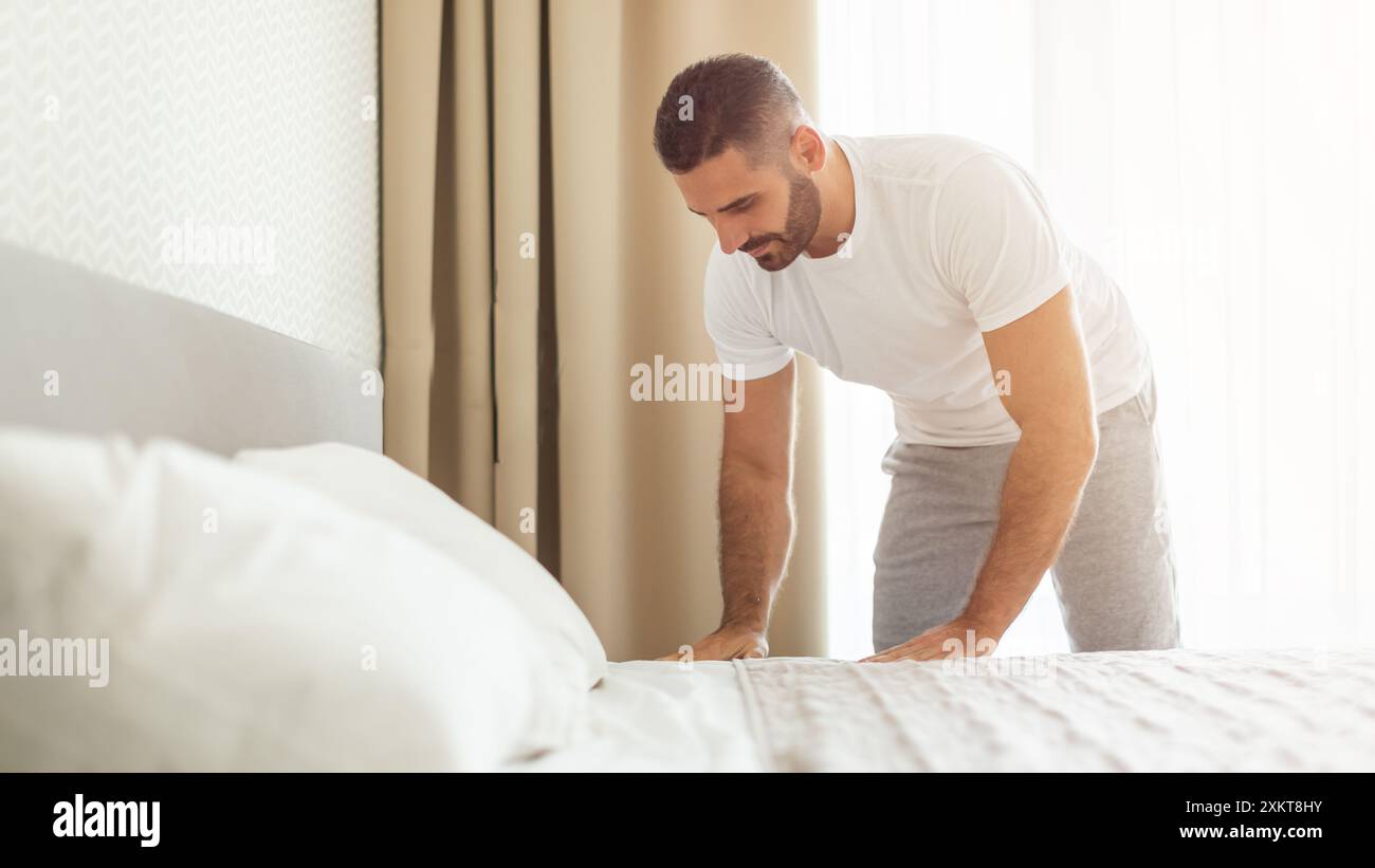 Ein Mann in weißem T-Shirt und grauer Hose beugt sich um, um sein Bett in einem hellen, modernen Schlafzimmer zu machen. Das Bett ist mit weißen Laken und Kissen gemacht, und das Stockfoto