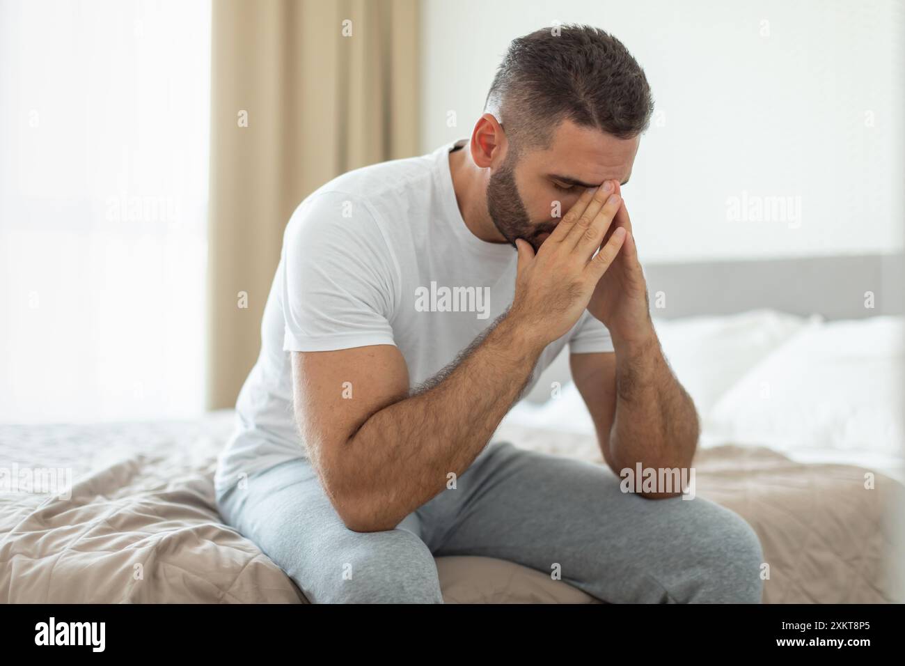 Ein Mann, der ein weißes T-Shirt und eine graue Hose trägt, sitzt am Rand eines Bettes. Er hat seine Hände im Gesicht und schaut nach unten, scheint in Dee zu sein Stockfoto