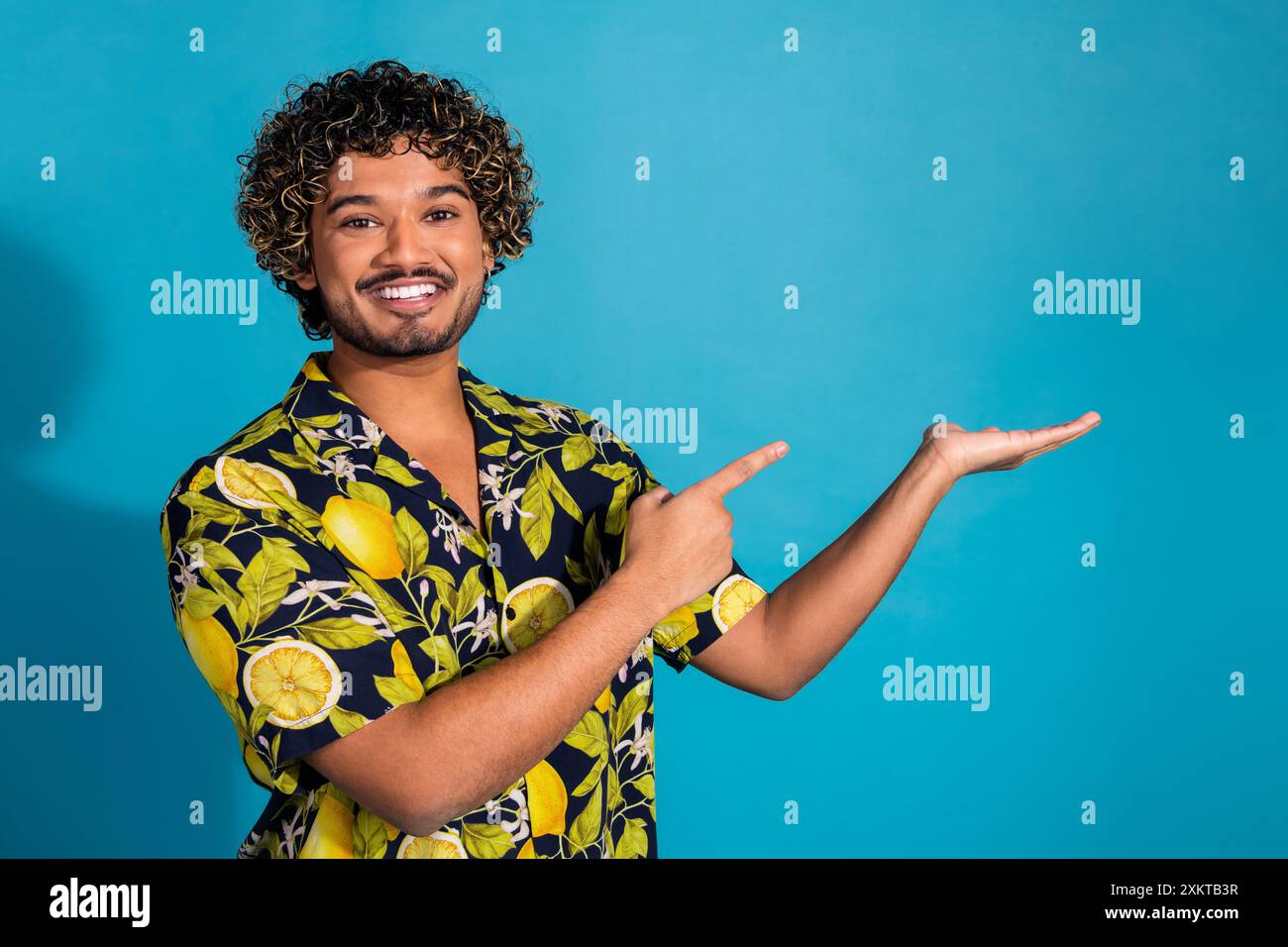 Foto eines guten Mannes mit gewelltem Haar gekleidetem Print-Hemd, das auf Objekt auf Arm leeren Raum lokalisiert auf blauem Hintergrund zeigt Stockfoto