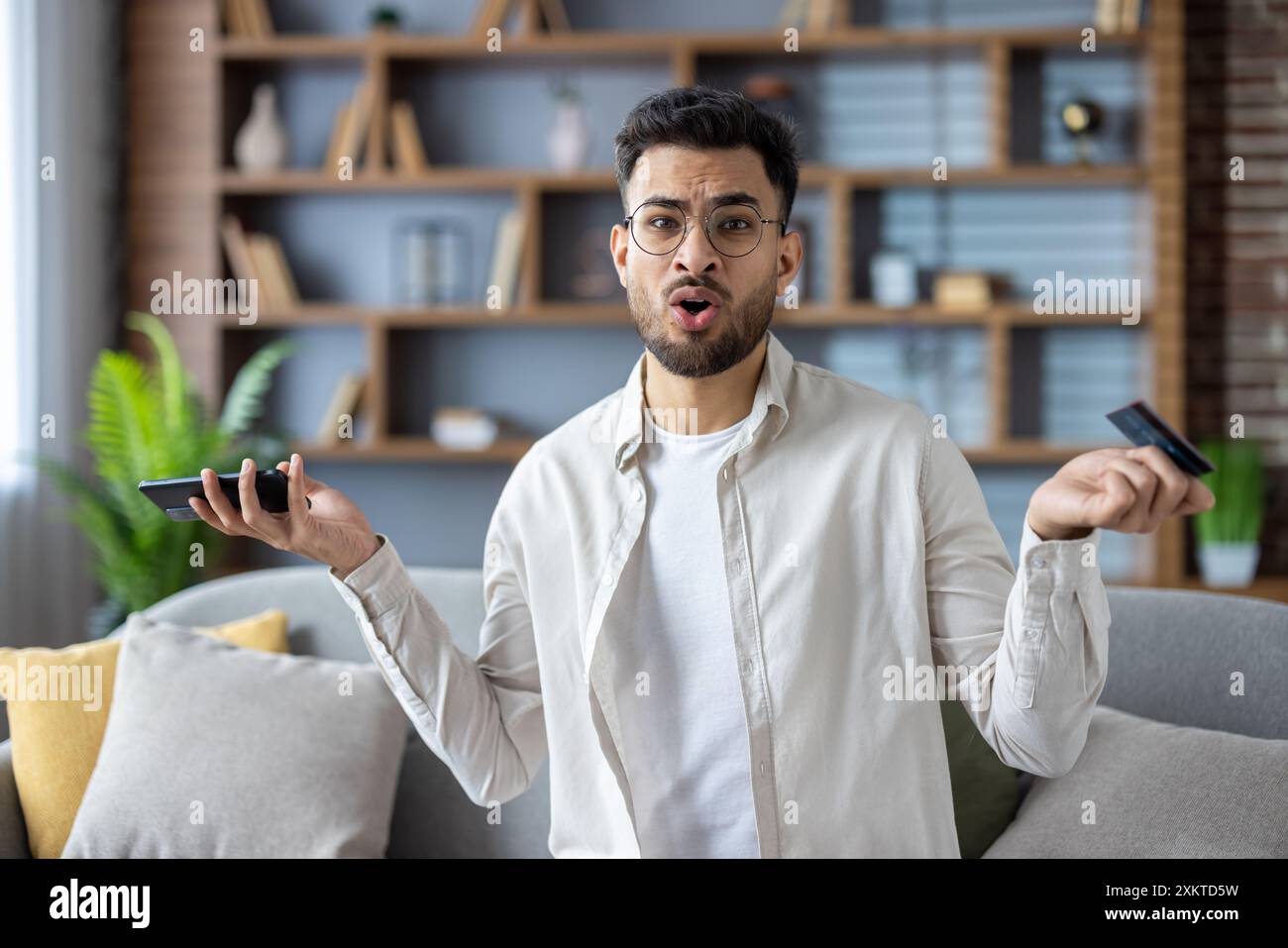 Junger Mann mit Brille, der auf dem Sofa sitzt und Smartphone und Kreditkarte in den Händen hält. Verwirrter Ausdruck, der die finanzielle Situation oder das Online-Shopping-Problem im modernen Wohnzimmer hinterfragt. Stockfoto