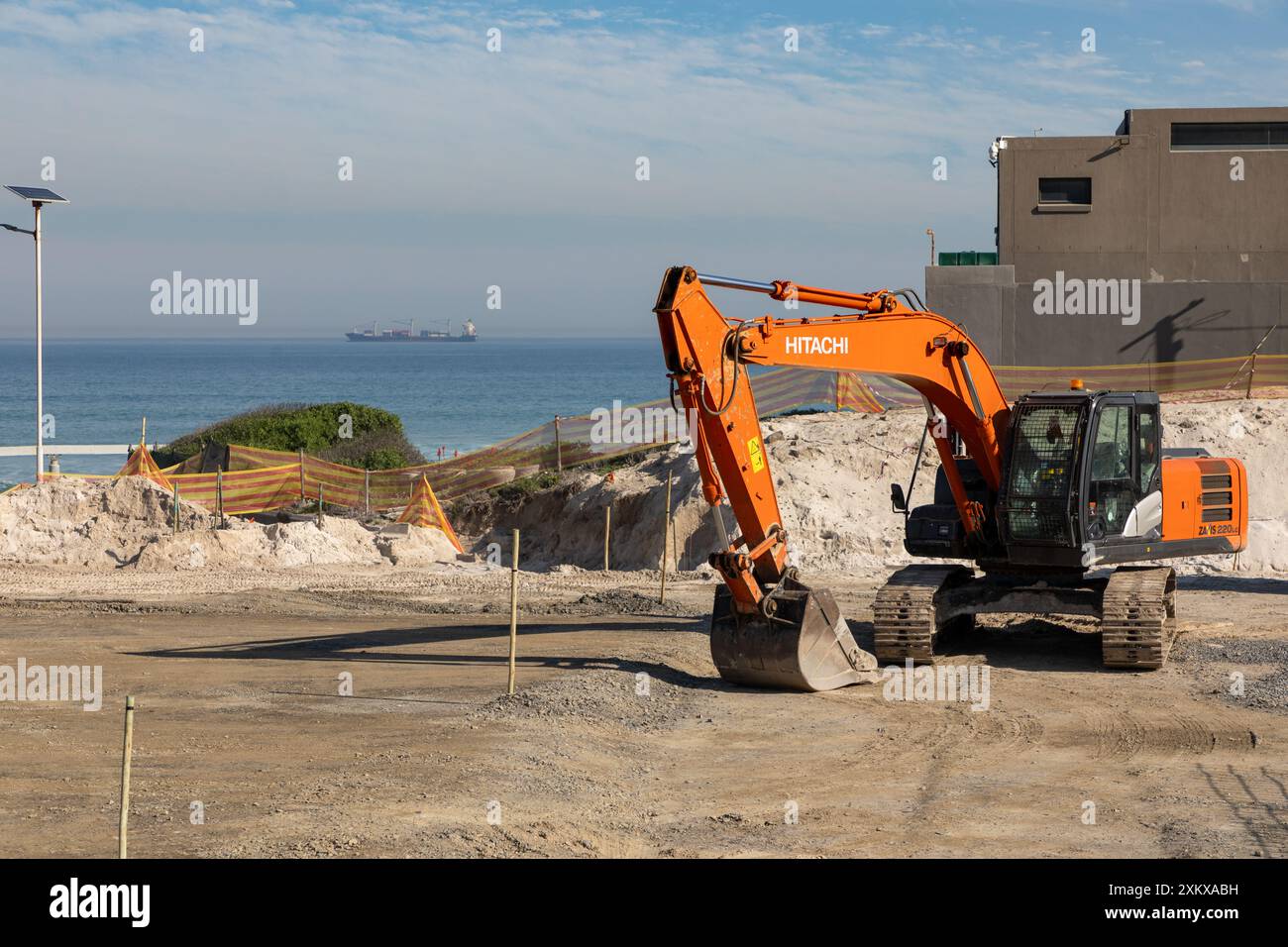 Hitachi Raupenbagger auf einer Baustelle mit dem Atlantik und einem Containerschiff im Hintergrund. Stockfoto